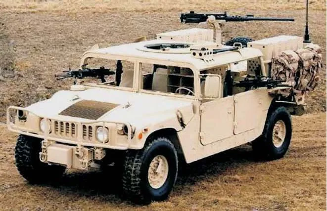A tan military high-mobility wheeled vehicle with roof-mounted and side-mounted machine guns, photographed on open grassland