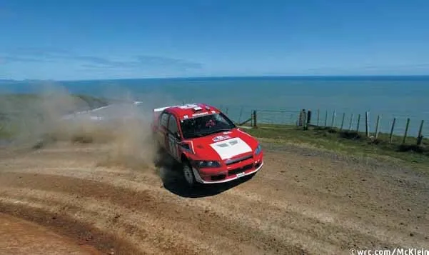 A rally car sliding through a gravel corner on a coastal road, its wheels turned into the slide and a plume of dust thrown up behind it, with the sea in the background
