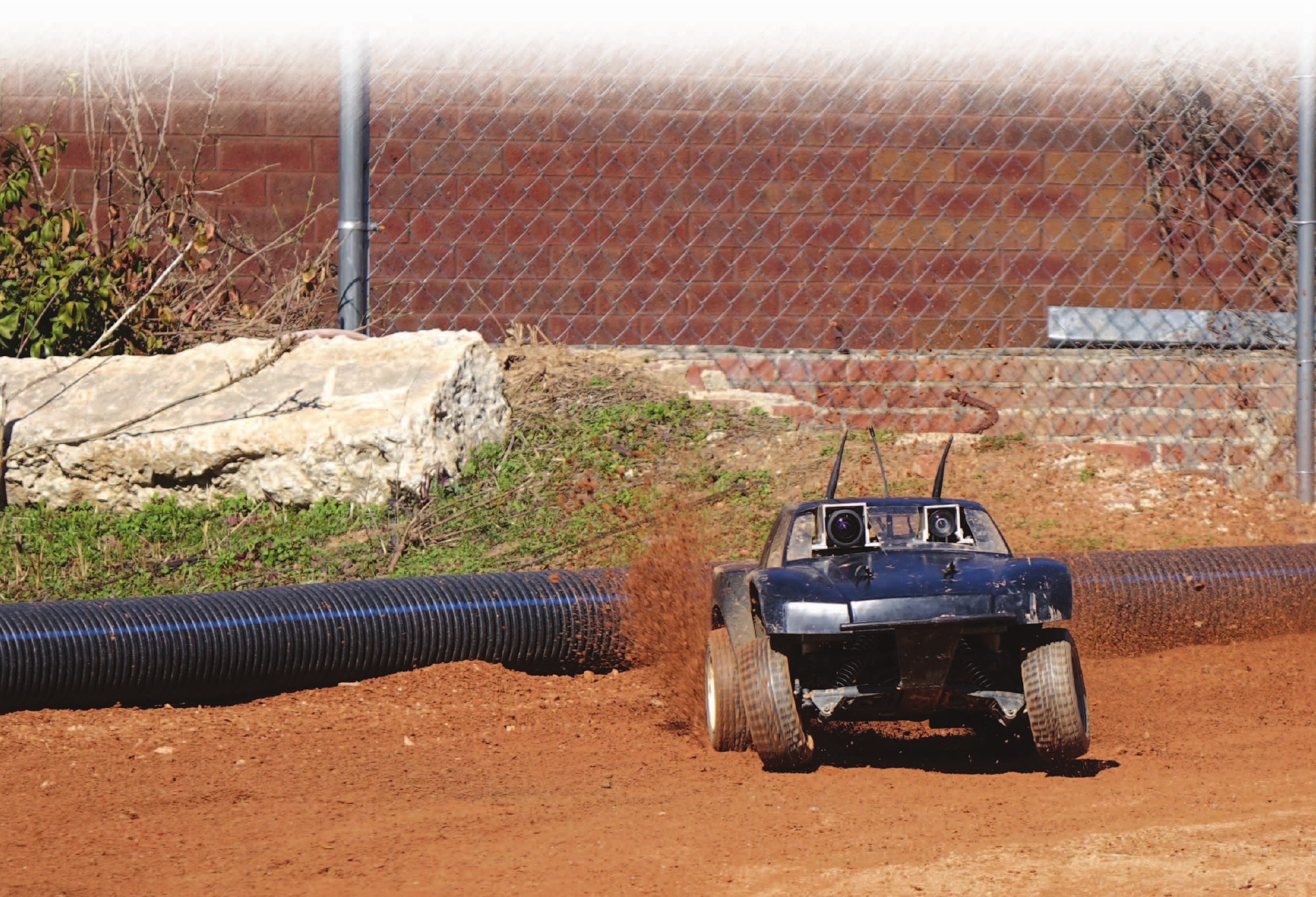 An AutoRally vehicle driving fast on a red dirt track, throwing up dust, with a stereo camera pair mounted above the body shell