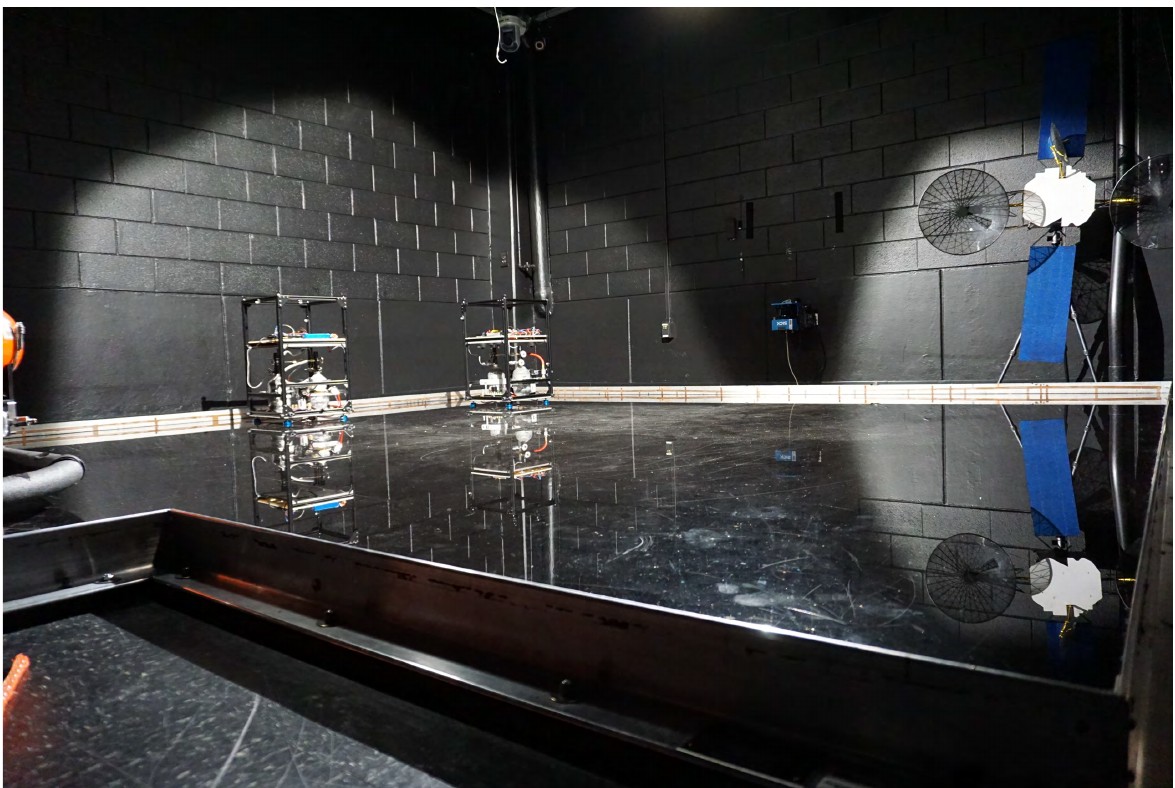 The darkened test arena: two COSMOS sleds stand on a black polished floor that reflects them, with a model satellite mounted on the far wall and spotlights picking out the scene